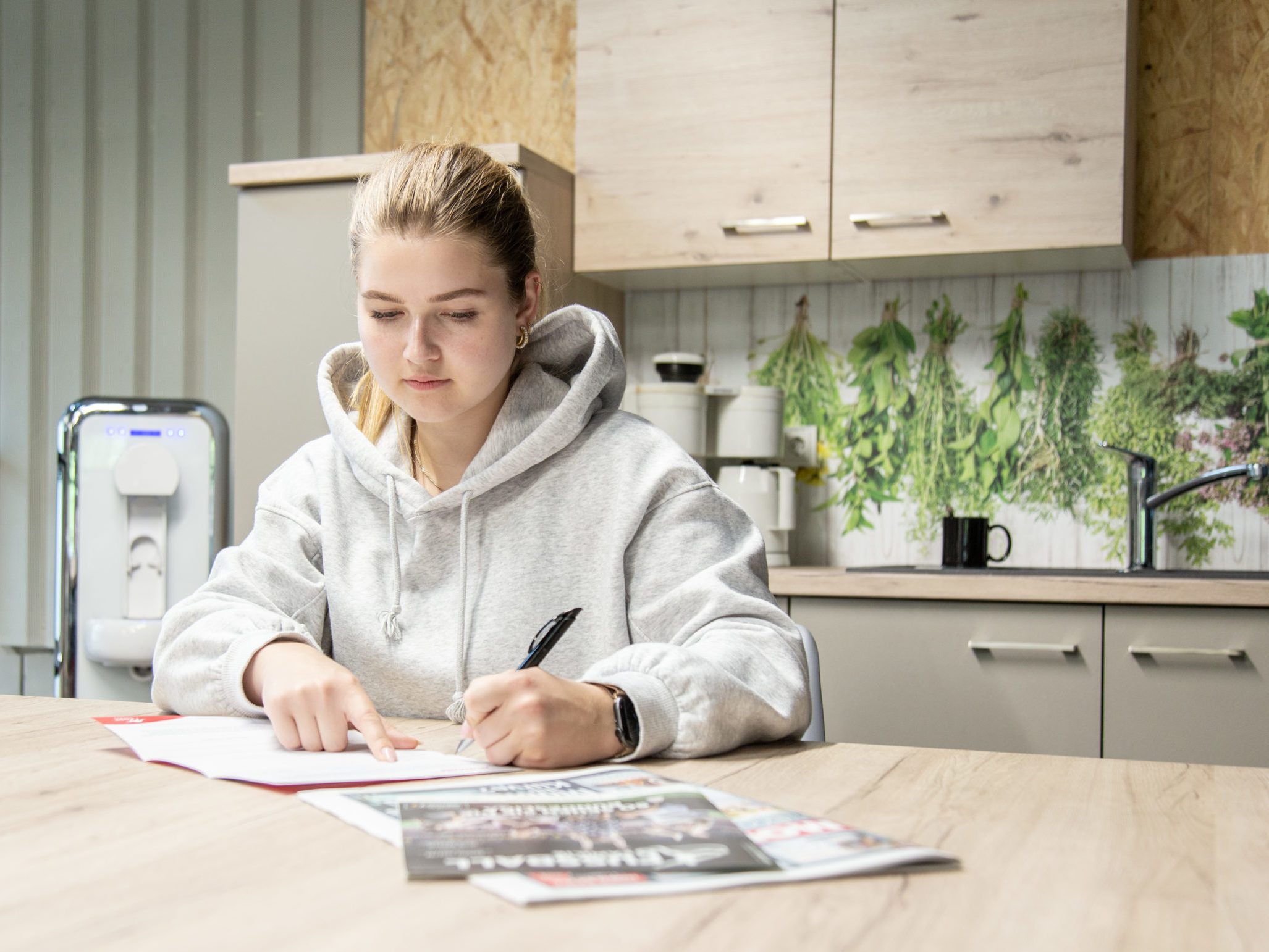 Das Bild zeigt eine junge Frau in einer Küche. Sie schreibt mit einem Kugelschreiber auf einem Blatt Papier von MediaLogistik. Das Bild nimmt Bezug auf Tipps und Tricks zur Bewerbung.